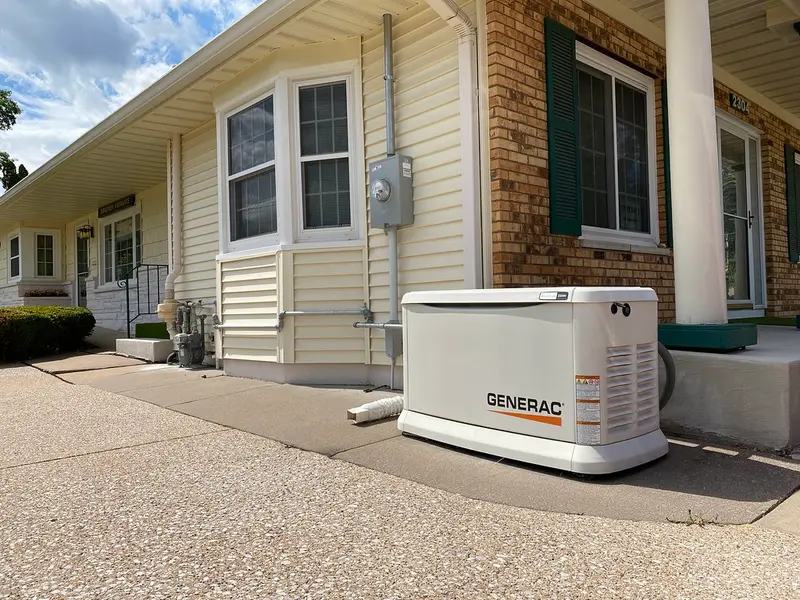 A white Generac standby generator installed on the ground beside a building, wired to gas piping and an electric meter on the wall