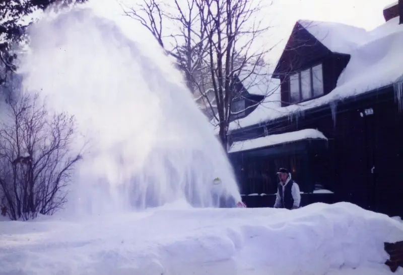 A person snow-blowing next to a house, throwing a tall arc of snow into the air