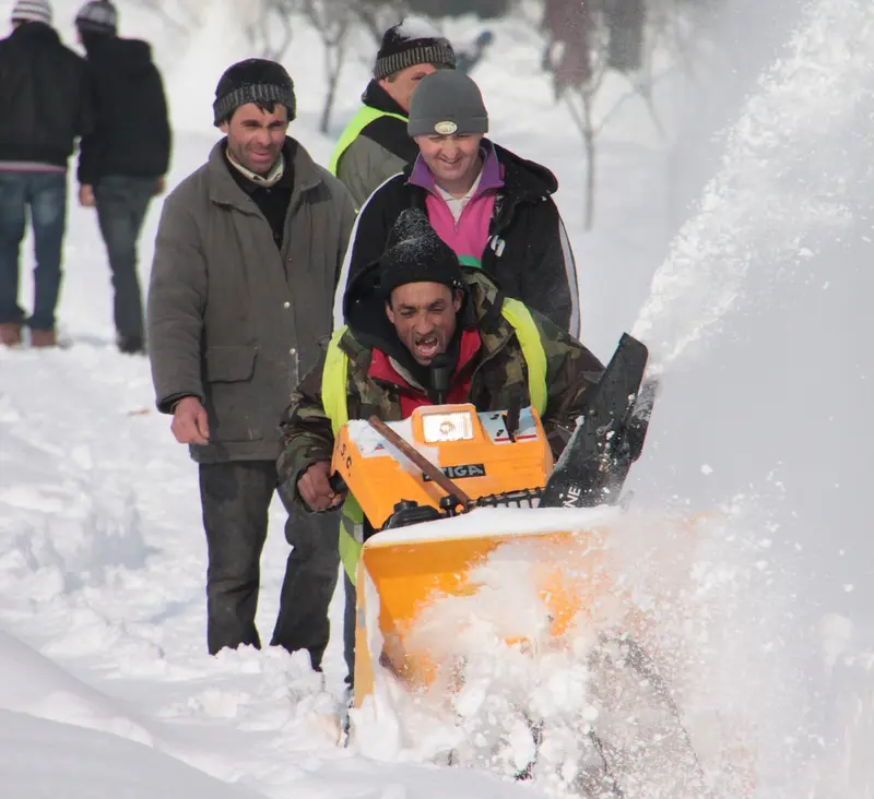 A man operating an orange walk-behind snow thrower in deep snow as other people look on